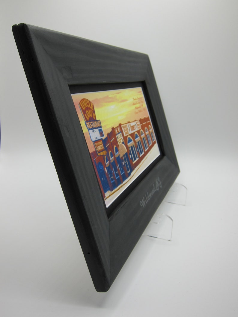 May include: A black wooden frame with a vintage postcard inside. The postcard shows a building with the text "Uncle Lou's Restaurant" on the front. The frame is on a white surface with two clear acrylic stands.