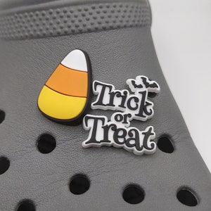 May include: A gray shoe with a candy corn charm and a "Trick or Treat" charm. The candy corn charm is white, orange, and yellow. The "Trick or Treat" charm is white with black lettering and a bat design.