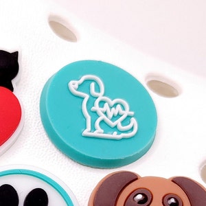 May include: A teal blue round charm with a white outline of a dog with a heart and EKG symbol inside.