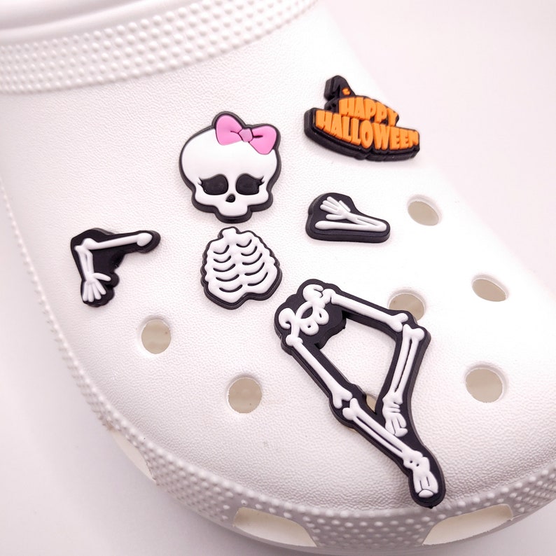 May include: A set of Halloween-themed shoe charms featuring a skeleton, a skull with a pink bow, a witch's hat, and the words "Happy Halloween".