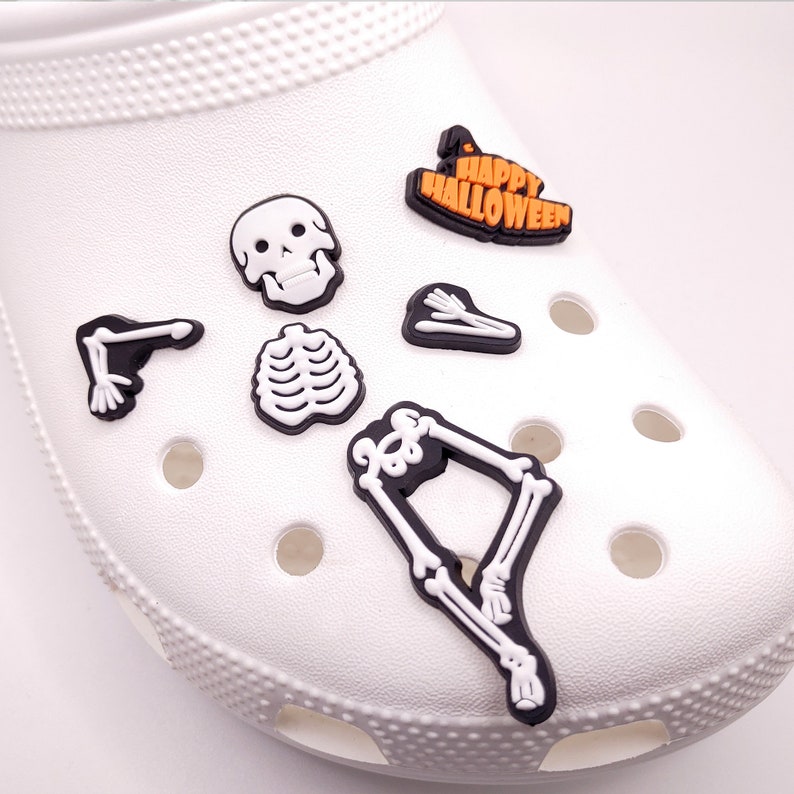 May include: White shoe with various Halloween themed shoe charms. The charms include a skull, a ribcage, a skeleton arm, a skeleton leg, and a charm that says "Happy Halloween".