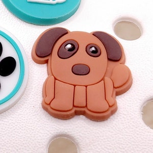 May include: A brown cartoon dog charm with brown ears, a brown nose, and black eyes. The charm is made of rubber and has a flat back.