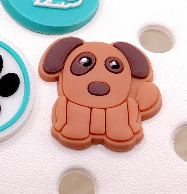 May include: A brown cartoon dog charm with brown ears, a brown nose, and black eyes. The charm is made of rubber and has a flat back.