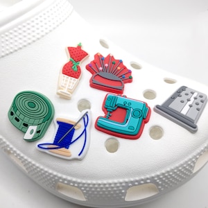 May include: Seven colorful sewing-themed shoe charms. The charms include a spool of thread, a sewing machine, a thimble, a pincushion, a dress form, and two spools of thread.
