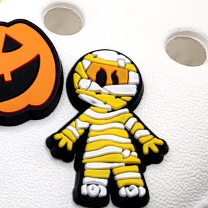 May include: A yellow and white mummy costume charm with black outlines. The charm is a cartoon-style mummy with a white bandage wrapped around its head and body. The charm is a Halloween-themed decoration.