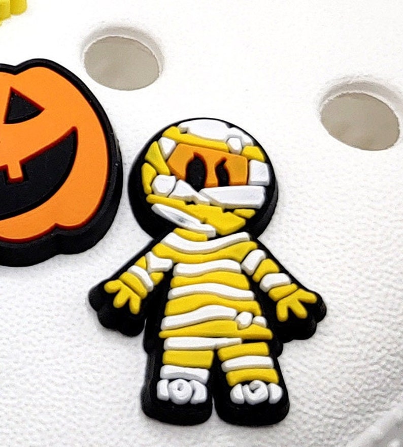 May include: A yellow and white mummy costume charm with black outlines. The charm is a cartoon-style mummy with a white bandage wrapped around its head and body. The charm is a Halloween-themed decoration.