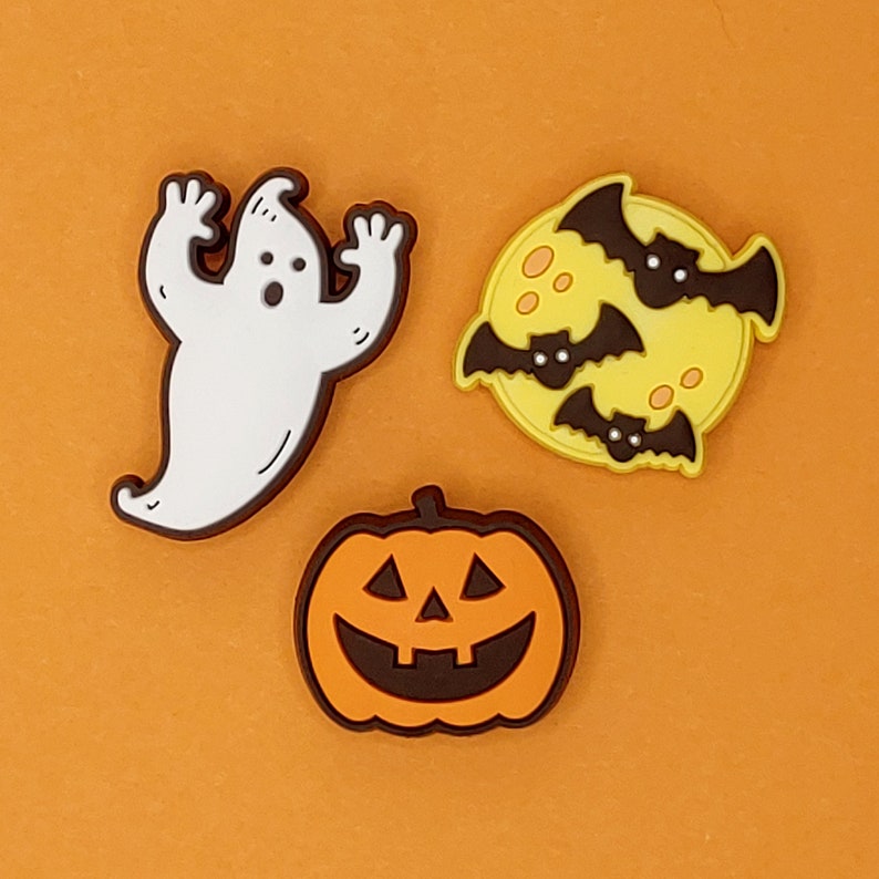 May include: Three Halloween-themed rubber charms: a white ghost, a jack-o'-lantern with a toothy grin, and a yellow moon with three black bats flying around it.