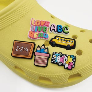 May include: A yellow shoe adorned with colorful, school-themed charms. Charms include the words "LOVE LIVE READ", "ABC", a yellow school bus, a chalkboard with "2+2=4", a pencil holder, and a charm that says "Teach".