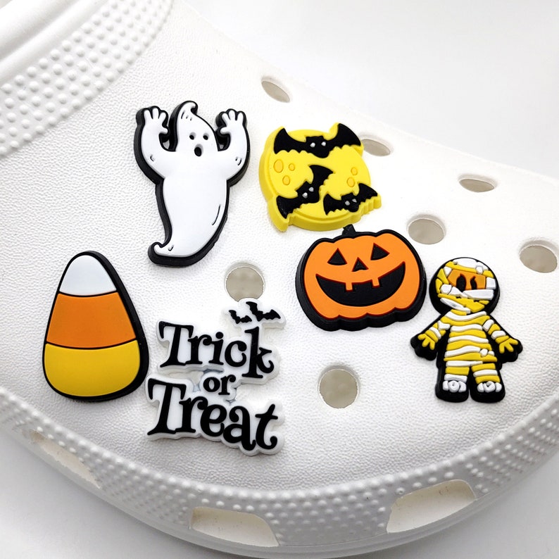May include: A white shoe with Halloween-themed shoe charms. The charms include a ghost, a pumpkin, a candy corn, a bat, and a "Trick or Treat" charm.