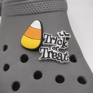 K&ouml;nnte beinhalten: Zwei wei&szlig;e und schwarze Halloween-Schuhcharms. Ein Charm ist ein Maisbonbon und der andere Charm sagt "Trick or Treat" mit einem kleinen Fledermausdesign.