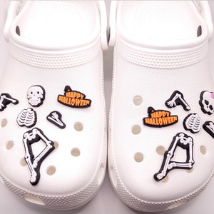 May include: A pair of white clogs with Halloween-themed shoe charms. The charms include a skeleton, a skull, a ribcage, a witch's hat, and the words "Happy Halloween".