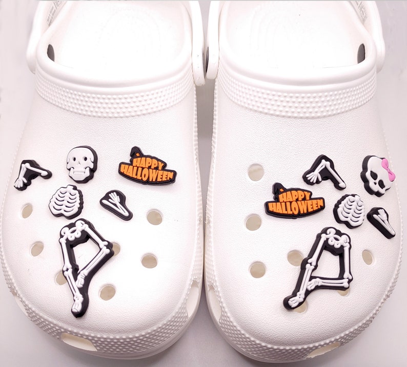 May include: A pair of white clogs with Halloween-themed shoe charms. The charms include a skeleton, a skull, a ribcage, a witch's hat, and the words "Happy Halloween".