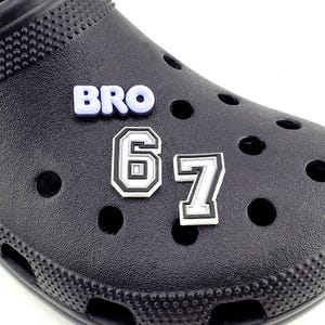 May include: Black shoe with decorative charms. The charms include the word "BRO" in light blue letters and the numbers "67" in a white and black design. The shoe has multiple holes and a textured surface.