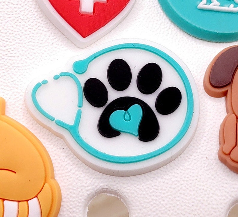 May include: A white charm with a turquoise outline featuring a black paw print with a turquoise heart inside. The charm is shaped like a stethoscope.