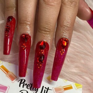 K&ouml;nnte beinhalten: Lange, rote, glitzernde Nagelspitzen mit Strasssteinen. Die N&auml;gel befinden sich auf einer Hand mit einem rosa, flauschigen Hintergrund. Der Text "Pretty Lit" ist in der rechten unteren Ecke des Bildes sichtbar.