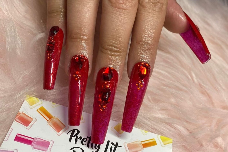 K&ouml;nnte beinhalten: Lange, rote, glitzernde Nagelspitzen mit Strasssteinen. Die N&auml;gel befinden sich auf einer Hand mit einem rosa, flauschigen Hintergrund. Der Text "Pretty Lit" ist in der rechten unteren Ecke des Bildes sichtbar.