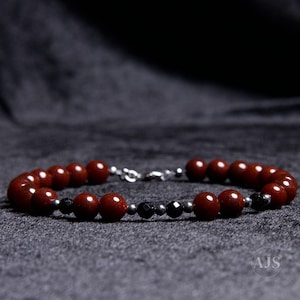 May include: A beaded bracelet featuring round, reddish-brown stone beads interspersed with smaller, faceted black beads and tiny metallic silver spacers. The bracelet is secured with a silver-toned metal lobster claw clasp and jump ring closure.