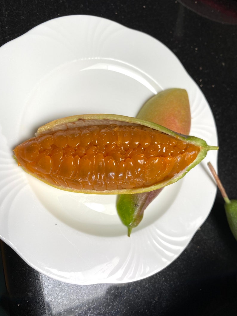 May include: A halved, open fruit with a vibrant orange interior, presented on a white plate. The fruit's outer shell is green and partially peeled back, revealing the juicy, segmented flesh. Two whole fruits are also visible.