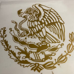 May include: Gold-colored decal featuring the Mexican coat of arms. The design includes an eagle with a snake in its beak, perched atop a cactus, surrounded by a wreath. The emblem is a symbol of Mexican national identity.