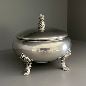 May include: A silver sugar bowl with a pineapple finial on the lid and three ornate feet. The bowl is sitting on a grey surface.