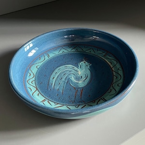 May include: A round, ceramic bowl with a blue glaze. The interior features a stylized bird design in shades of blue and white, surrounded by a decorative border. The bowl's rim is a lighter shade of blue.