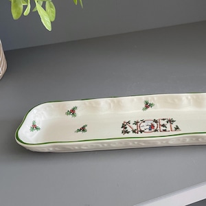 Puede incluir: Una bandeja de servicio rectangular de cerámica color hueso con bordes festoneados y un borde verde. El interior presenta un patrón repetitivo de pequeñas hojas de acebo y bayas rojas, con la palabra NOEL centrada en letras decorativas.