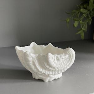 May include: A white ceramic bowl shaped like a seashell with a glossy finish. The exterior features intricate, raised ridges and textures that mimic the natural form of a mollusk shell, while the interior is smooth and deep with a scalloped rim.