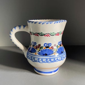 Könnte beinhalten: Eine weiße Keramikkanne mit einem blauen, grünen und roten Blumendesign. Die Kanne hat einen Henkel und einen runden Körper.