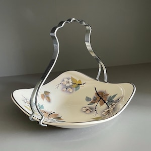 May include: Cream-colored, vintage serving dish with a silver handle. The dish features a floral and leaf design in muted tones of brown, yellow, and blue. The handle is arched and has a decorative, wavy top.