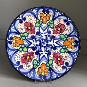Könnte beinhalten: Ein runder Keramikteller mit einem handbemalten Blumenmuster in leuchtendem Rot, Orange und Grün auf weißem Grund. Das Design zeigt komplexe blaue Schnörkel, schraffierte Details und einen durchgehenden schwarzen Außenrand.