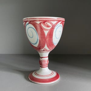 May include: A white ceramic goblet with a red and blue painted design. The goblet has a wide bowl, a slender stem, and a circular base. The bowl features red swirls and blue accents. The base has red stripes.