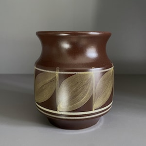 May include: A brown ceramic vase with a wide, rounded body and a slightly flared rim. The vase features a repeating pattern of stylized, light-colored leaf shapes. The design is accented with white stripes.