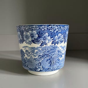 Puede incluir: Una maceta de cerámica con un patrón de transferencia en azul y blanco. El diseño muestra un paisaje detallado con árboles, figuras y un castillo, rematado por un borde floral decorativo. El recipiente tiene forma cilíndrica y base blanca.