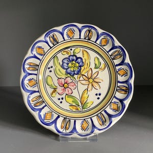 Könnte beinhalten: Ein dekorativer Keramikteller mit gewelltem Rand. Der Teller hat ein Blumendesign mit blauen, rosa und gelben Blumen. Der Rand ist weiß mit blauen und orangefarbenen Akzenten. Der Teller wird auf einem transparenten Ständer präsentiert.