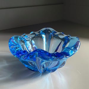 Puede incluir: Un cuenco de cristal azul transparente con un borde festoneado. El cuenco tiene forma de flor con un color azul oscuro alrededor de los bordes que se desvanece a un azul más claro en el centro. El cuenco está sobre una superficie blanca.