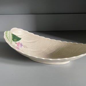 以下が含まれることがあります： クリーム色の葉の形をしたセラミック製サービングディッシュ。この皿は、テクスチャ加工された表面を持ち、緑の葉とピンクの花柄のアクセントで装飾されています。この皿の長さは約30cmで、浅く湾曲したデザインです。