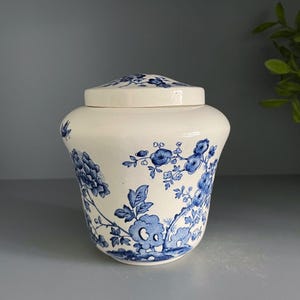 May include: A cream-colored ceramic jar with a matching lid, featuring a blue floral pattern of blossoms and branches. The jar has a rounded body and a smooth finish, with the blue botanical design wrapping around the exterior surface.
