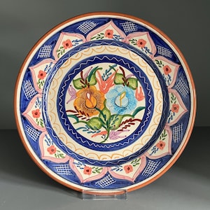 May include: A round ceramic plate featuring a hand-painted floral design in the center with orange and blue blossoms. The border displays a repeating pattern of pink petals, blue geometric cross-hatching, and small red flowers on a cream base.