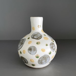 May include: A small, white ceramic vase with a rounded body and a narrow neck. The vase is decorated with a pattern of gray, yellow, and green spots. The vase is approximately 6 inches tall.