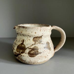 May include: A speckled, off-white ceramic pitcher with a rounded body and a curved handle. Dark brown leaf-like patterns are painted on the pitcher's surface. The pitcher has a small spout for pouring.