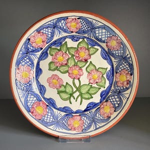 May include: A decorative ceramic plate with a floral design. The plate features a central arrangement of pink flowers with yellow centers and green leaves. The rim is orange, and the border has blue and white patterns.