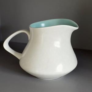 Puede incluir: Una jarra de cerámica blanca con un borde azul claro. La jarra tiene un cuerpo redondeado y un asa.