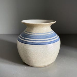以下が含まれることがあります： 手作りの陶器の花瓶で、まだらなベージュのボディと水平な青いストライプがあります。花瓶は丸みを帯びた形状で、わずかにフレアしたリムが付いています。この装飾品は、花を飾ったり、単独のアートオブジェクトとして使用するのに適しています。