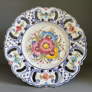 May include: A round ceramic plate with a scalloped edge and intricate cut-out details. The center features a hand-painted floral arrangement in pink, yellow, and blue. The border is decorated with blue scrollwork and small pink and blue flowers.