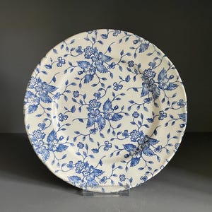May include: A round ceramic plate featuring an off-white base color with a repeating blue floral pattern. The design consists of intricate blue flowers, leaves, and thin winding vines covering the entire surface of the plate in a dense arrangement.