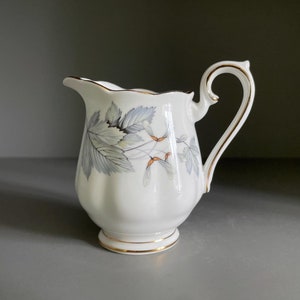 May include: A white ceramic creamer with a gold trim and a floral design featuring gray leaves and brown accents.