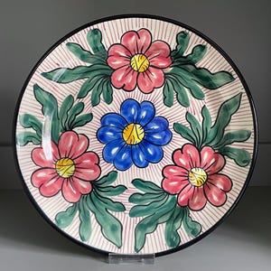 May include: A round ceramic plate with a black rim, featuring a floral design. The plate has a cream background with pink and white stripes. The flowers are pink and blue with yellow centres, surrounded by green leaves.