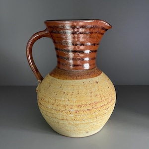 May include: A handmade ceramic pitcher with a rounded base and a handle. The pitcher has a speckled, light tan body and a dark brown, striped top. The spout is on the top of the pitcher.