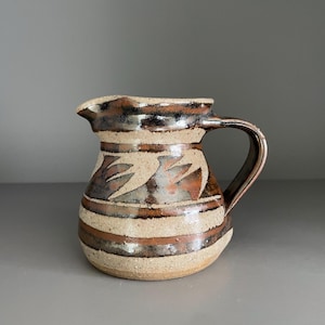 May include: A handmade ceramic pitcher with a dark brown handle and spout. The pitcher features horizontal bands of tan and brown, with a decorative pattern of stylized leaves. The pitcher is a rustic, handcrafted piece.