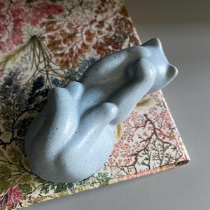May include: A pale blue ceramic cat ornament curled up on a patterned fabric. The fabric features a floral and tree design in pink, green, and brown hues. The background is a light grey surface.